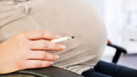 A close up of a pregnant woman's belly. She is sat in a black chair and is wearing a beige top and blue jeans and is holding a cigarette in her hand. 