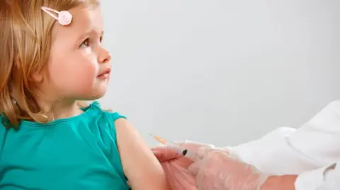 Getty Images a small girl wearing a green t-shirt with one sleeve rolled back looks at the person next to her. Only the arms of the other person can be seen, wearing a white shirt or coat, clear plastic medical gloves, holding a syringe next to the child's arm, ready to give a vaccine