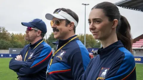 Apple TV Three people are seen dressed in blue sports gear. The man on the left has a blue baseball gap and sunglasses, the man in the middle has a white visor, sunglasses and moustache, and on the end is a woman with brown hair tied back. They are standing in a football stadium