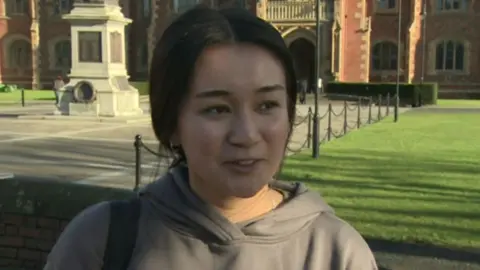 A student with brown hair outside QUB Lanyon Building. There is grass behind. She is wearing a brown hoody.