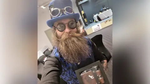 Lewis Aylmer taking a selfie, holding a box of a flask and two shot glasses. He is wearing a black, long-sleeved shirt, a blue and black waistcoat, black glasses and a blue top hat. He has a long, bushy, brown beard and a long, brown moustache. 
