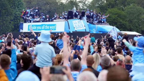 A crowd of people wearing predominately sky blue watch players and staff of Coventry City parade in a sky blue-coloured bus. One player in black holds aloft the Championship trophy.