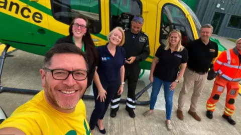 Mike Land Mike Land in a bright yellow t-shirt taking a selfie with several members of the Wiltshire Air Ambulance, all in uniform, and standing in front of an air ambulance