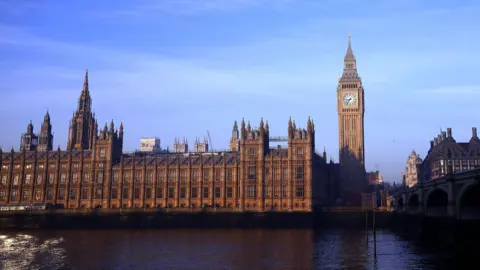 The Houses of Parliament line the river Thames to the left with the Big Ben structure on the right, below a blue sky on a bright day. The glow of the sun is making the gothic brick buildings appear a dark orange.