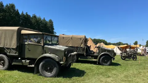 Two Army green, historical trucks are parked alongside each other on the grass in a field. A historical motorbike in the same colouring is parked behind them. 