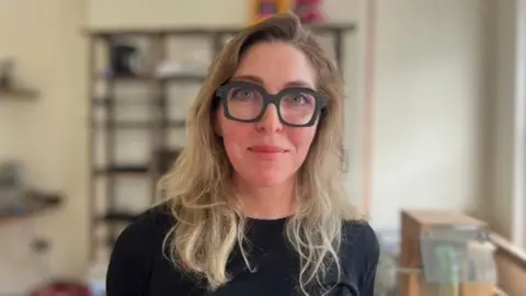 Claire Mulvenna stands in her opticians shop. She is wearing thick black rimmed glasses, and a a black jumper. She has long blond hair.