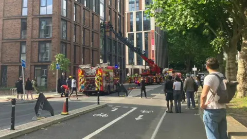 Firefighters at a scene outside a block of flats. People can be seen walking down a fire engine ladder and onlookers stand in the street facing the flats