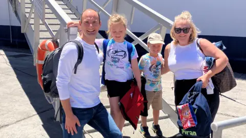 A family, of a husband and wife, and two children, a girl and a boy, about to board the ship The Global Mercy. They are holding bags and luggage. 