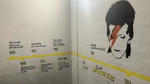 A horizontal yellow line is painted on a brick wall, which is painted white. Against it are various years with brief notes, starting on the left, from 1842: "Corn Exchange opens, built on St Andrew's Hill" to 1968. The entry for 1966 says: "David Bowie plays with his band The Buzz" and there is a stencilled image of Bowie as Ziggy Stardust with a red lightning flash painted across his face.