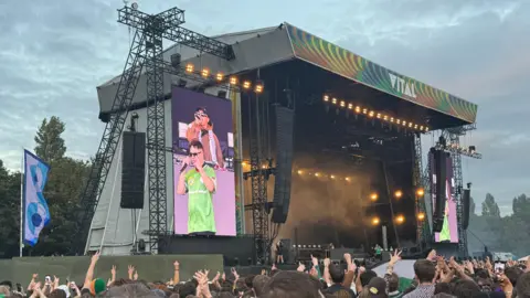 A large pop up stage outside. It has two speakers on either side and two large screens displaying images of Irish rap trio Kneecap who are performing. A large number of people are gathered for the gig and many of them are holding their hands in the air. 