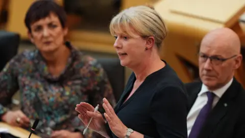 PA Media Shona Robison standing speaking in the Scottish Parliament. She is in the foreground, with her hands apart, holding a pen. Her hair is tied back and she is dressed in black. Seated behind her are Angela Constance, to the left, and John Swinney.