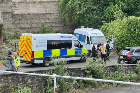 Two police vans are parked on the road outside the building, while people in high-vis jackets stand around.