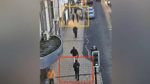 Crown Prosecution Service Still from CCTV footage showing a figure in black following behind a young woman walking along a street.