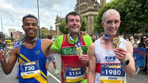 Three men in a line with running vests on, holding up their medals from the event and smiling at the camera