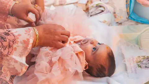 Sophia Kelemen lies on a changing mat. She is wearing a peach coloured outfit with ruffles and has a dummy in her mouth. A woman's hands can be seen adjusting her outfit.