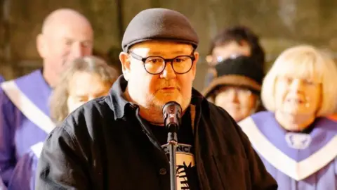 Laura Gates Comedian Johnny Vegas wearing a black cap, black glasses, black shirt and black and white t-shirt stands in front of a microphone with people blurred out in the background dressed in purple.
