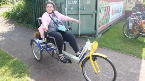 Milestones Trust A lady wearing a pink cardigan, black leggings and a bicycle helmet, sitting on an adapted trike on a footpath, with a rack of other bikes in the background