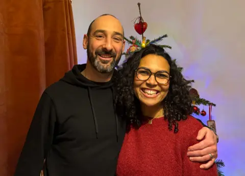 Daniel in black hoodie with his arm around Rachel in red jumper an glasses. Both are smiling and a Christmas tree is behind them