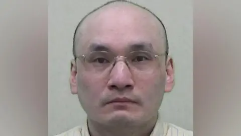 Northumbria Police Mugshot of Thomas Kwan, he is bald and wearing glasses and a yellow shirt with blue pinstripes.