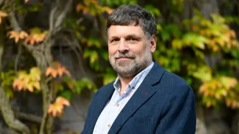 University of Aberdeen A man - Neil Curtis, head of collections at the University of Aberdeen - with a greying beard and wearing a dark blue suit jacket oven a light blue open-necked short, with green and yellow leaves on tree branches in the background. 