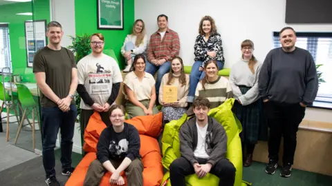 A team of mainly young people sit and stand in an office. They are smiling and a woman in the centre is holding an engraved wooden award. The front two people are sitting in large colourful beanbag chairs.