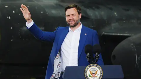 PA Media JD Vance stands waving at a lectern in front of a black US military plane in a hangar at RAF Fairford - he wears a blue suit jacket and white shire. The lectern has two microphones and the emblem of the US Vice President's Office