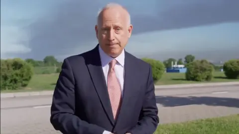 Steve Rosenberg standing outside in front of a plume of smoke in a screenshot taken from a TV report. He wears a navy suit and pink tie.