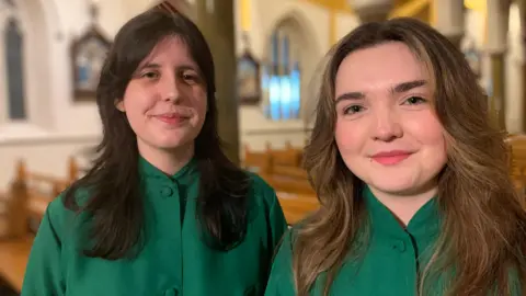 Lucy and Rebekah inside St Peter's Cathedral. They both have brown hair that is shoulder-length. Lucy on the left has darker hair. They are both wearing green dresses.