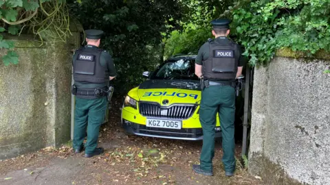 BBC Two uniformed police officers stand either side of a marked police car. It's parked at the end of a gated driveway.