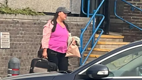 Rachel Goodman walks in front of the court, carrying a suitcase and wearing a cap which partially hides her face. She has long, straight, dark hair and wears a pink top and black trousers or leggings.