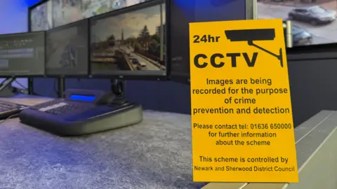 NEWARK AND SHERWOOD DISTRICT COUNCIL A bright yellow sign with a diagram of a CCTV camera and black writing on a desk