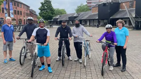 Seven people are standing on a pavement, many of them are wearing helmets and holding bikes. 