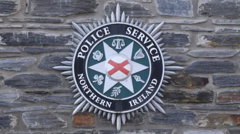 On a grey stone wall hangs a PSNI badge. The badge is silver black and green