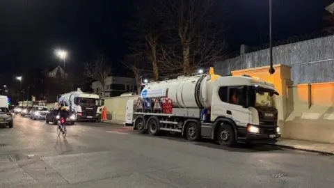 A Thames Water tanker lorry parked on a residential street at night, with traffic queuing behind it and a cyclist riding past. Streetlights illuminate nearby houses and a concrete wall on the right.