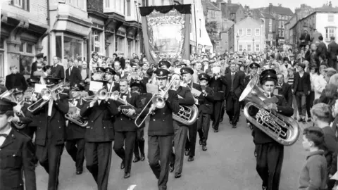 Getty Images A black and white picture of a large crowd of people parading down a street, which is lined with shop fronts. The group in front are a brass band, wearing matching suits, hats, and playing various instruments. A banner is being held up behind them. There are people watching from the sides.