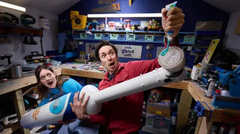 PA Ruth and Shawn in their workspace, holding the toothbrush as Shawn squirts toothpaste onto its head. Both are wearing amazed expressions.