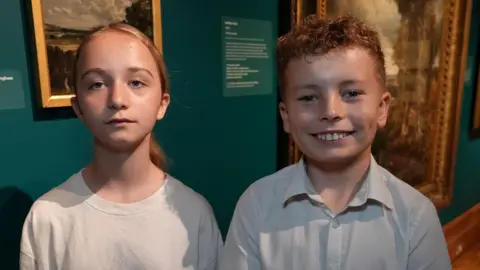 Qays Najm/BBC A young school girl and boy smile at the camera in an art exhibition. The girl has blonde hair that has been tied back and is wearing a white T-shirt. The boy has curly short red hair and is wearing a white school shirt.