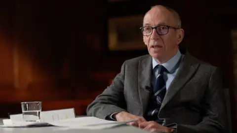 Sir Rob Behrens, who has receding grey hair and glasses, and wears a grey wool suit over a blue shirt and tie, sits at a desk in front of some documents and a glass of water. 