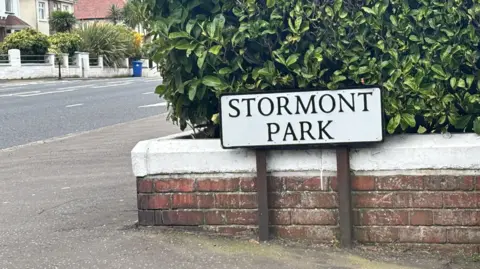 A low-level street sign for Stormont Park. The immediate background is a low brick wall and some bushes. Houses can be seen off to the left.