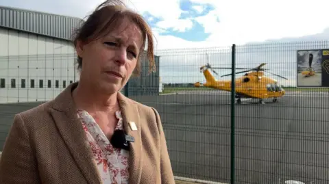 Karen Jobling is wearing a beige coat with a floral blouse. Her head is tilted to the right. She has brown hair. Behind her is a yellow helicopter.
