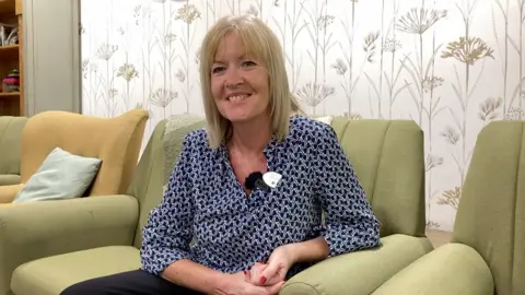 A blonde lady in a blue and white geometric blouse with nails painted red smiles at the camera. She sits on a green chair while a small black microphone is clipped to the neck of her blouse.