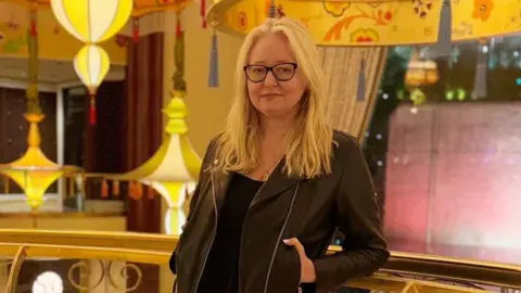 A woman with long blonde hair and black-rimmed glasses stands in front of a railing with lights and bright decorations in the background. She is wearing a black top underneath what looks like a black leather jacket.