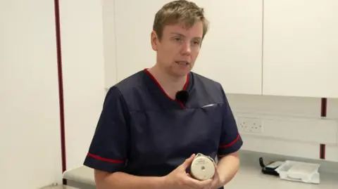 Nurse consultant Lindsey Pollard. She is wearing a dark blue short sleeve nursing top edged with red around the sleeves and neck. She has short, light brown hair. She is holding a round silvery metal disc - the pain pump - which is about 2cm thick.