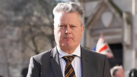 Vincent Kearney, a blue-eyed, grey-haired man in a grey suit, white shirt and orange and black striped tie. Behind him blurred is a man holding a Union Jack flag and a grey stone building. 