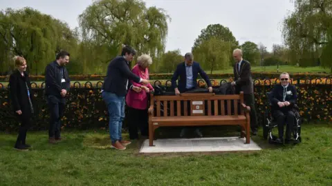Legend on the Bench A group of seven people stood on a grassy verge around a bench.