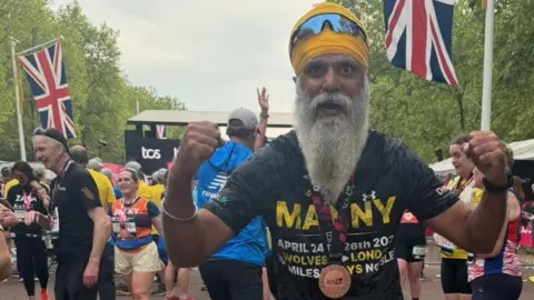 A man with a grey beard is wearing a black T-shirt including the word Manny in yellow and other words is holding out both fists and looking at the camera. Other people are behind him and the union jack flags are also visible.