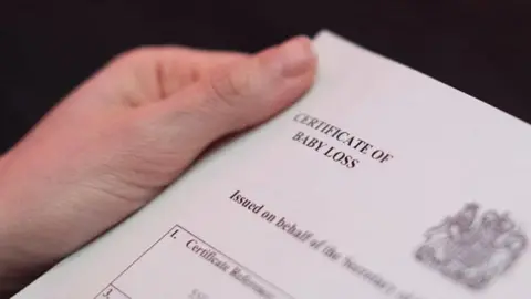 An image of a certificate of baby loss, just the top left corner of the paper is visible, and a hand is holding it out to read. 