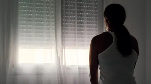 Getty Images A woman sits on her bed facing the window. The blinds are nearly all the way down. Her hair is tied back and she's wearing a white vest top.