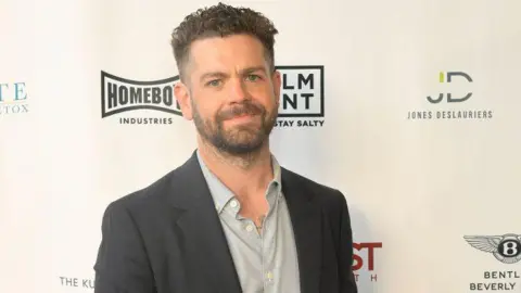 Jack Osbourne, who has dark hair cropped short and a beard, standing in front of a hoarding bearing various company logos. He is wearing a grey suit jacket and pale grey shirt and smiling.