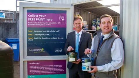 Lincolnshire County Council The paint shop is in a shipping container with shelves of different kinds of paints.  The two councillors stand at the entrance, each with a tin of paint in their hands.